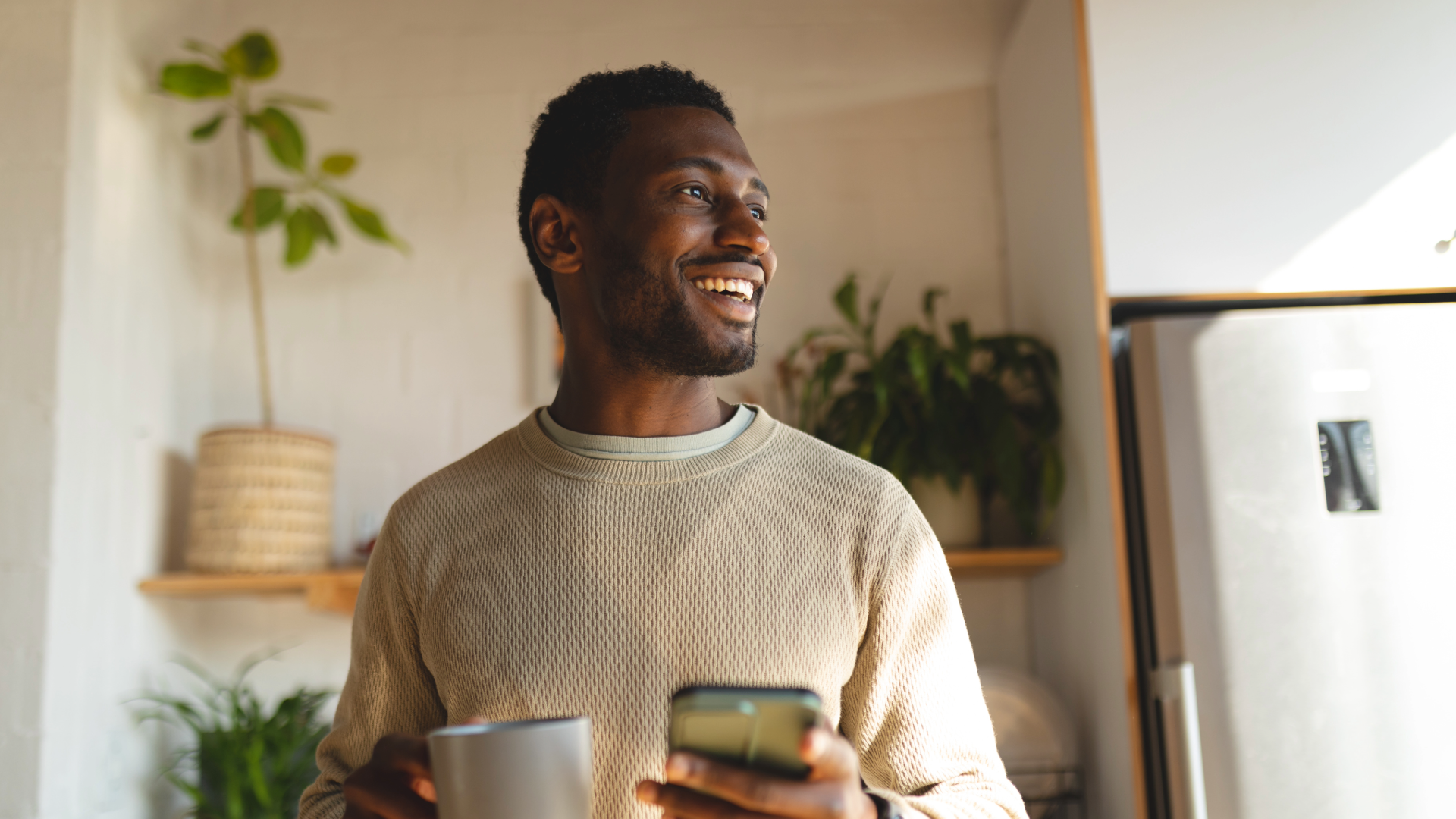 Man holding a mug and phone, smiling in a home setting with plants and a refrigerator. Single Origin Club.
